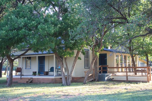 A cozy, light-colored house surrounded by trees, featuring a porch and a grassy yard.