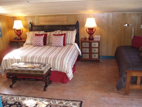 Cozy bedroom with a striped bed, two lamps, a nightstand, and a small seating area in a rustic setting.