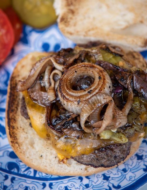 Close-up of a juicy burger with melted cheese, grilled onions, and peppers on a toasted bun, served on a decorative plate.