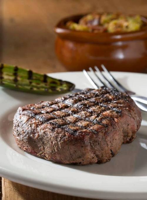 A perfectly grilled steak on a white plate, with a side of grilled asparagus and a bowl of vegetables in the background.