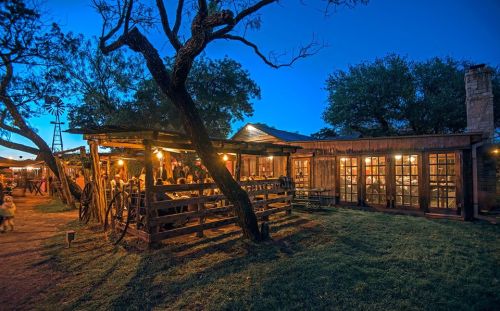A rustic outdoor dining area with string lights, surrounded by trees, at dusk. A cozy atmosphere with people gathered.