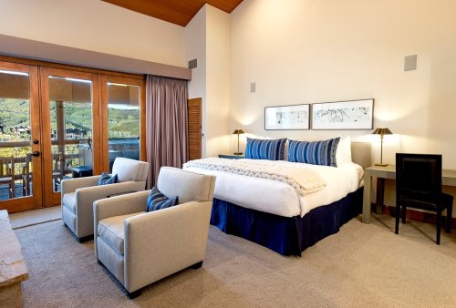 Cozy hotel room with a large bed, two armchairs, and a balcony overlooking a scenic view. Warm, inviting decor.