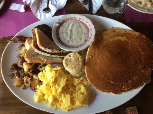 A hearty breakfast plate featuring scrambled eggs, pancakes, sausage, toast, biscuits, and a bowl of gravy.