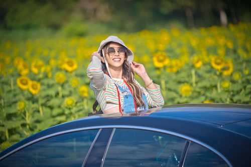 A woman in sunglasses and a hat smiles from a car, surrounded by a field of sunflowers.