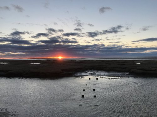 A serene sunset over a calm marsh, with clouds reflecting colors in the water and silhouettes of posts in the foreground.