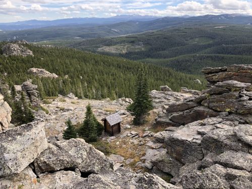A small wooden cabin nestled among rocky terrain and pine trees, overlooking a vast green valley and distant mountains.