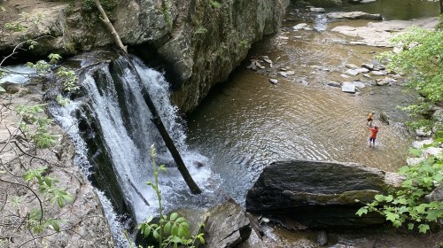 A waterfall cascades into a rocky stream, with two people standing in the water nearby, surrounded by lush greenery.