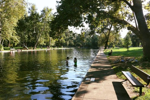 Two children play in a calm pond, surrounded by trees and people enjoying a sunny day in a park.