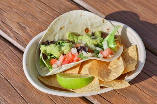 A taco filled with guacamole, lettuce, and tomatoes, served with tortilla chips and a lime wedge on a wooden table.