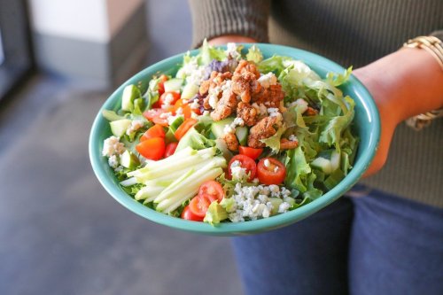 A person holding a vibrant salad bowl filled with greens, cherry tomatoes, apples, and crunchy toppings.