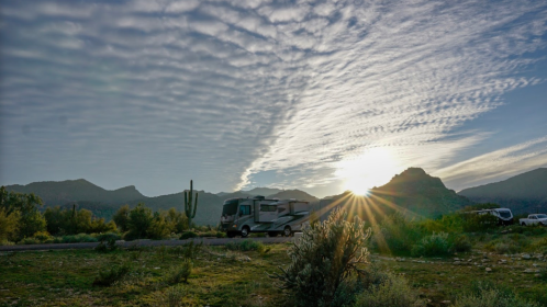 An RV parked in a scenic landscape at sunrise, with mountains and clouds in the background.