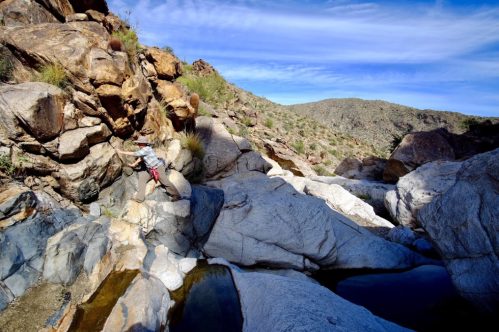 A person climbing rocky terrain in a desert landscape with blue skies and scattered clouds.
