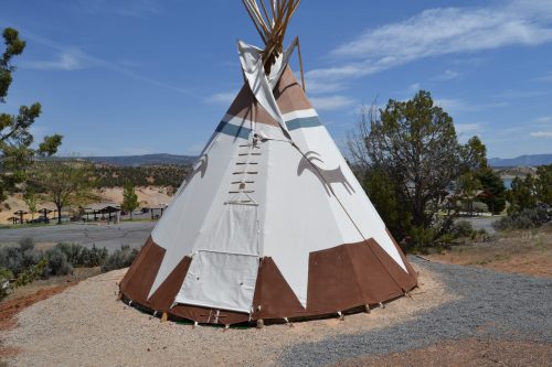 A traditional teepee with a painted design, set against a scenic landscape with trees and mountains in the background.