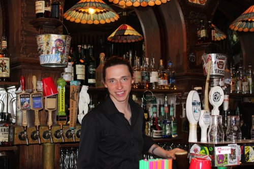 A smiling bartender stands behind a bar filled with colorful bottles and taps, surrounded by vintage decor.