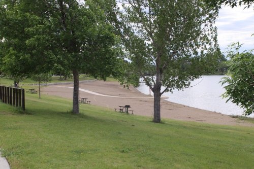 A serene park scene featuring trees, a sandy beach, and a calm lake under a cloudy sky. Picnic tables are visible.