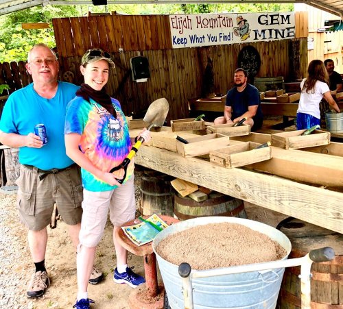 A woman with a shovel smiles at a gem mining site, while a man stands nearby holding a drink. Others are in the background.