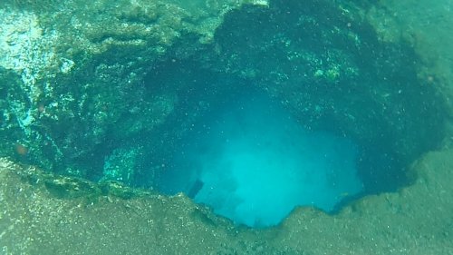 Underwater view of a rocky cave opening, revealing clear blue water inside.