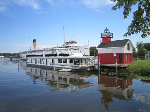 A red lighthouse beside a docked boat, with a large ship in the background, reflecting on calm water under a blue sky.