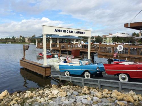A dock labeled "Amphicar Launch" with colorful amphibious cars parked by the water under a cloudy sky.