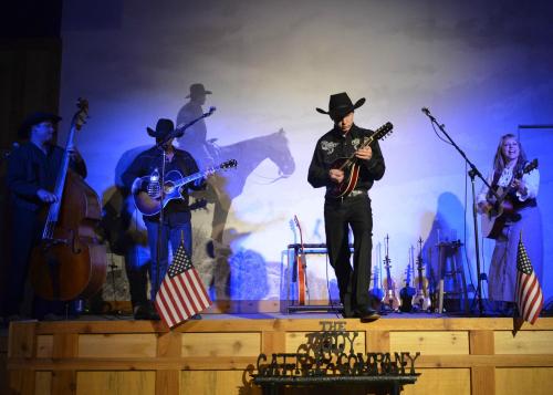 A country band performs on stage with instruments, American flags, and a cowboy silhouette in the background.