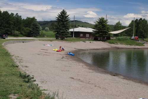 A sandy beach by a calm lake, with trees in the background and people relaxing on colorful floats.