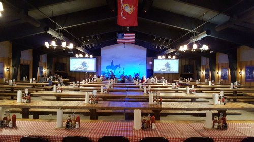 A spacious dining hall with wooden tables, red checkered tablecloths, and a stage featuring a band, decorated with flags.
