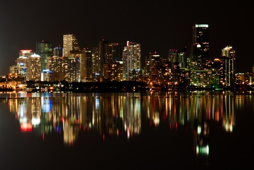 City skyline at night, illuminated buildings reflecting on calm water.