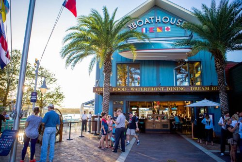 A vibrant waterfront restaurant with palm trees, people mingling outside, and flags waving in the breeze.