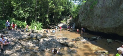 A scenic riverbank with people enjoying the water, surrounded by lush greenery and rocky cliffs on a sunny day.