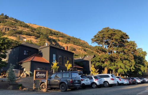 A modern building with a sign reading "Society" surrounded by trees and parked cars, set against a hillside.