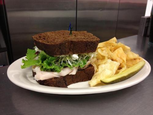 A sandwich with turkey, lettuce, and sprouts on dark bread, served with potato chips and a pickle on the side.