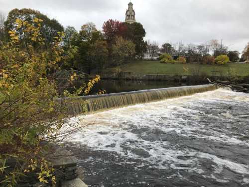 A waterfall cascades over a dam, surrounded by autumn foliage and a distant clock tower under a cloudy sky.