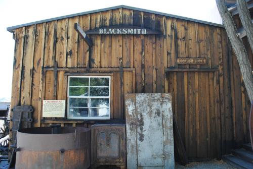 A rustic wooden blacksmith shop with a sign, window, and metal door, surrounded by gravel and trees.