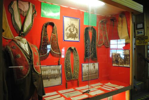 Display case featuring historical garments, photographs, and documents on a red background, showcasing traditional attire.