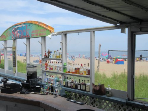 A beach bar with a colorful sign, showcasing various liquor bottles, overlooking a sandy beach filled with umbrellas and people.