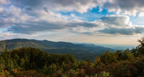A panoramic view of rolling mountains under a cloudy sky, with lush green trees in the foreground.