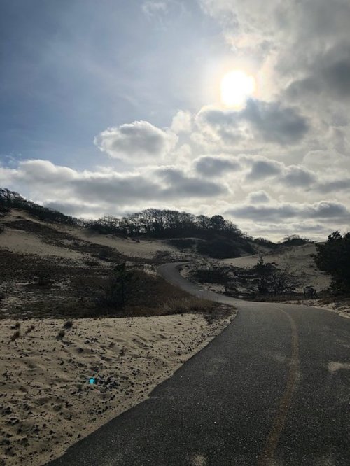 A winding path through sandy dunes under a cloudy sky with the sun peeking through.