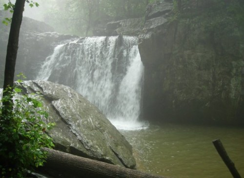 A serene waterfall cascades over rocks into a misty pool, surrounded by lush greenery.
