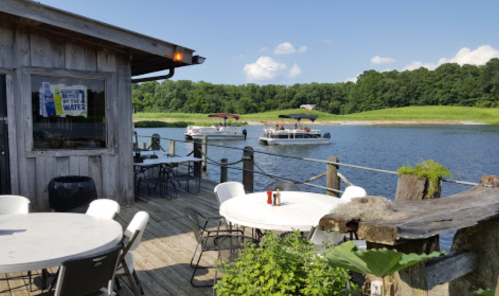 A wooden deck by a lake with tables, overlooking boats on the water and green hills in the background.