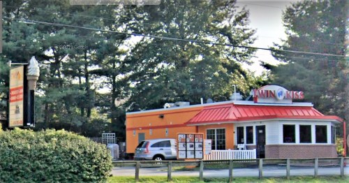 A colorful ice cream shop named "Twin Kiss" with a red roof, surrounded by trees and a sign in front.