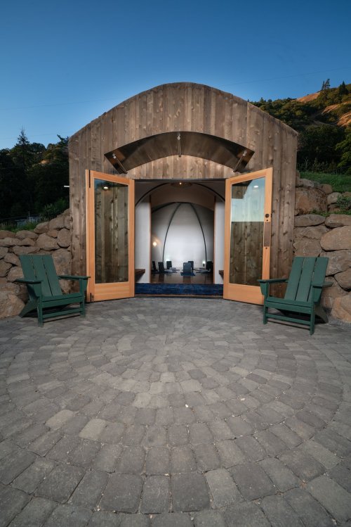 A modern wooden structure with open doors, surrounded by stone and featuring two green chairs in front.
