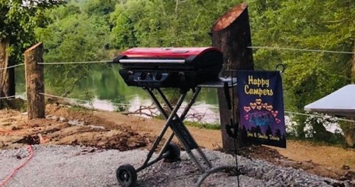 A red grill on a stand near a river, with a "Happy Campers" sign hanging nearby and trees in the background.