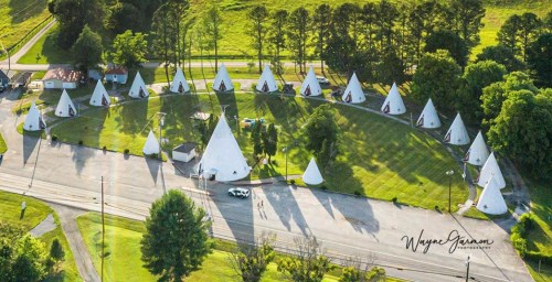 Aerial view of a circular arrangement of white teepee-shaped cabins surrounded by green grass and trees.