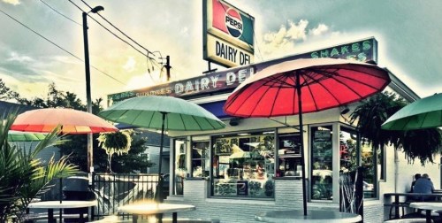 A colorful ice cream stand with umbrellas, featuring a Pepsi sign, surrounded by trees and a cloudy sky.