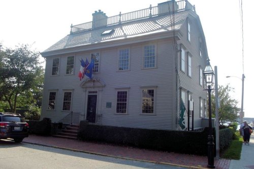 Historic gray house with a flag, surrounded by greenery and a brick walkway, featuring a porch and multiple windows.