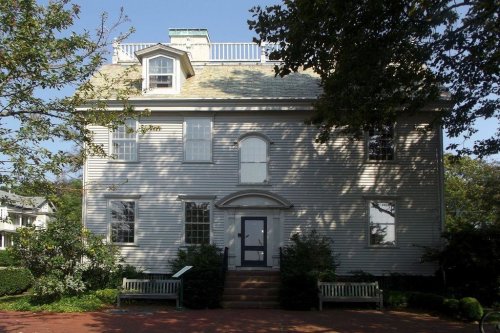 A large, two-story house with a gray exterior, white trim, and a small rooftop deck, surrounded by greenery.