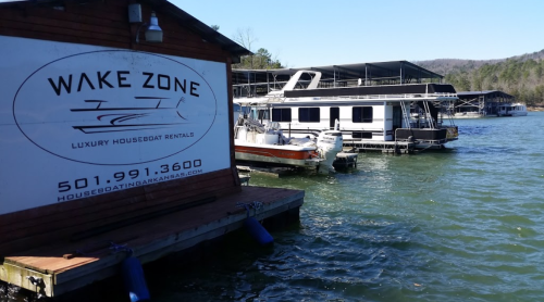 A dock with houseboat rentals on a lake, featuring a sign for "Wake Zone" and various boats moored nearby.