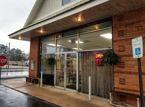 A brick building with large glass windows, an "OPEN" sign, and potted plants, set on a rainy day.
