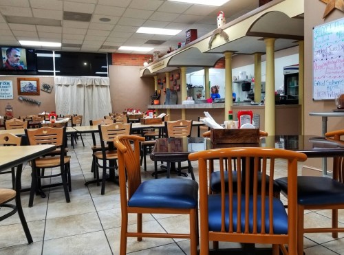 A cozy restaurant interior with empty tables, a counter, and a TV showing a program in the background.