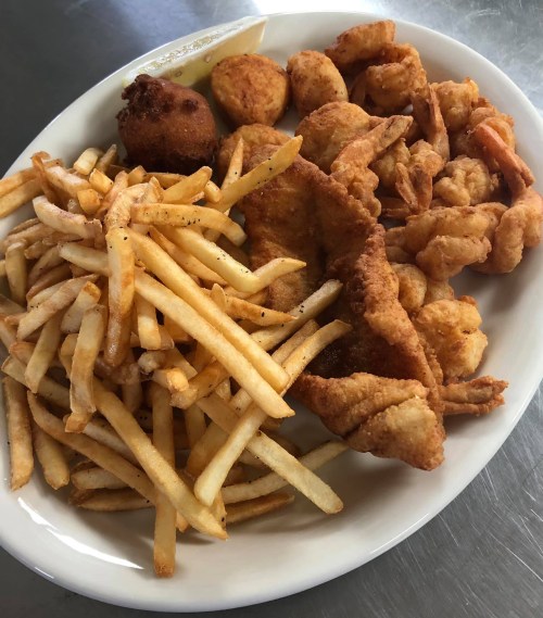 A plate of fried seafood including shrimp, fish, hushpuppies, and a generous serving of French fries.